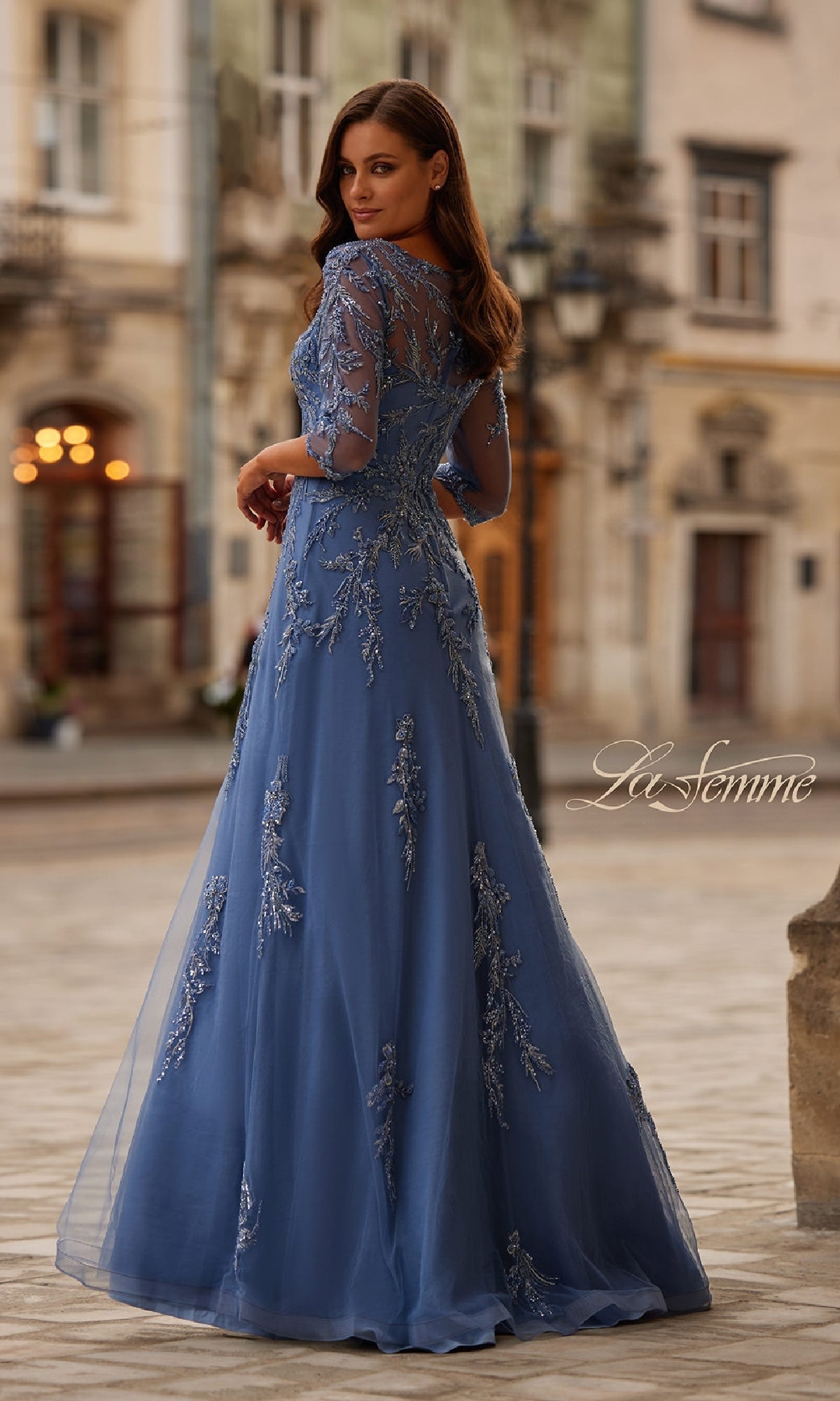 A woman stands outdoors in the La Femme 32612, a long blue beaded formal dress with sheer sleeves and floral embroidery—an elegant choice for a mother of the bride, set against a historic city square backdrop.